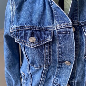 May include: A medium-wash denim jacket with a classic collar and silver-tone buttons. The jacket features a chest pocket with a button closure and detailed stitching. A timeless outerwear piece.