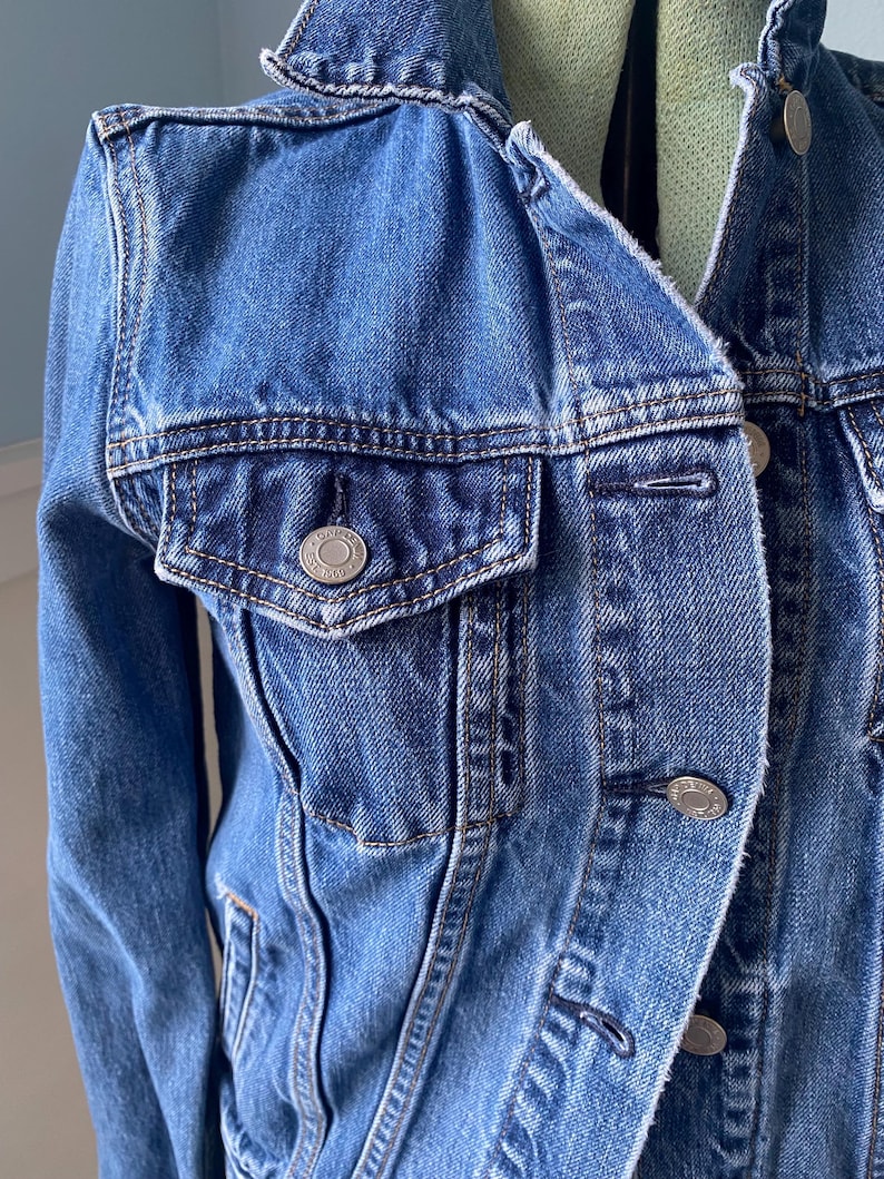 May include: A medium-wash denim jacket with a classic collar and silver-tone buttons. The jacket features a chest pocket with a button closure and detailed stitching. A timeless outerwear piece.