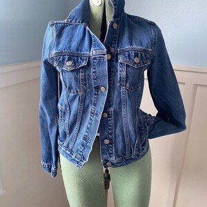 May include: A classic blue denim jacket with silver-tone buttons and two chest pockets. The jacket is displayed on a dress form. The jacket has a collared neckline and long sleeves. A timeless wardrobe staple.