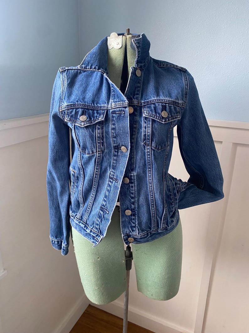May include: A classic blue denim jacket with silver-tone buttons and two chest pockets. The jacket is displayed on a dress form. The jacket has a collared neckline and long sleeves. A timeless wardrobe staple.