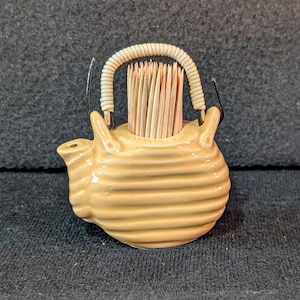 Może przedstawiać: Jasnożółty, żebrowany ceramiczny pojemnik na wykałaczki w kształcie czajniczka z zakrzywionym dzióbkiem i białym, teksturowanym, łukowatym uchwytem. Górny otwór wypełniony jest pękiem jasnobrązowych drewnianych wykałaczek ustawionych pionowo.