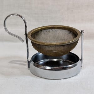 May include: A tea infuser with a metal frame and a mesh basket. The infuser has a handle and a small, round base to catch drips. The mesh basket is made of a fine, bronze-coloured material.