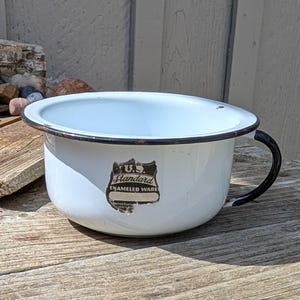 Può includere: Scodella in smalto bianco con bordo e manico neri. La scodella ha un adesivo bianco e nero con la scritta "U.S. Standard Enameled Ware".