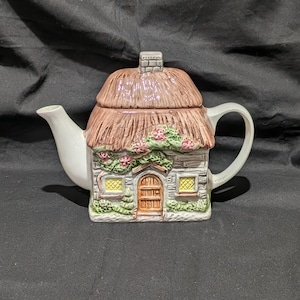 May include: A ceramic teapot shaped like a cottage. The roof is thatched, and the walls are stone-like with a door and windows. The teapot has a white spout and handle, with pink flowers and green vines. The chimney is gray.