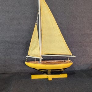 May include: A model sailboat featuring a yellow hull with a black lower section, two yellow fabric sails, and a white mast. The boat is mounted on a yellow cross-shaped wooden stand, showcasing a detailed deck with a hollow cockpit area.