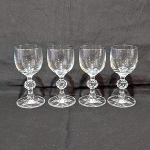 May include: Four clear crystal wine glasses with a decorative, faceted stem and a round base. The glasses are arranged in a row against a dark background. The glasses are likely used for serving wine or other beverages.