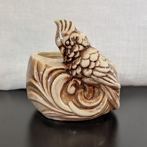 Pode incluir: Um vaso de cerâmica em forma de pássaro com esmalte marrom e branco. O pássaro está empoleirado em um design ondulado.