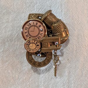 May include: A brass-toned brooch featuring a collection of miniature objects including two circular clock faces with Roman numerals, a sewing thimble, a small rectangular frame, and a circular gear component with a dangling key charm attached.