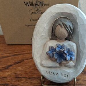 Puede incluir: Figurilla de cer&aacute;mica blanca de una mujer sosteniendo un ramo de flores azules. La figurita tiene forma ovalada y tiene las palabras "Gracias" inscritas en la parte inferior.