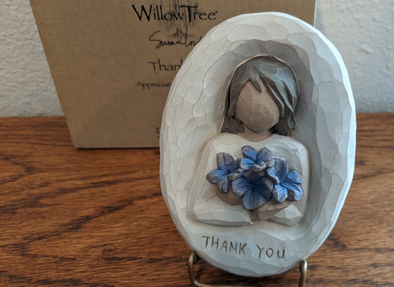 Puede incluir: Figurilla de cer&aacute;mica blanca de una mujer sosteniendo un ramo de flores azules. La figurita tiene forma ovalada y tiene las palabras "Gracias" inscritas en la parte inferior.