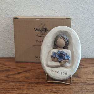 Puede incluir: Una figurita de madera tallada de una persona sosteniendo un ramo de flores azules. La figurita est&aacute; sobre un peque&ntilde;o soporte con las palabras "Gracias" inscritas en &eacute;l. La figurita est&aacute; hecha por Willow Tree by Susan Lordi.