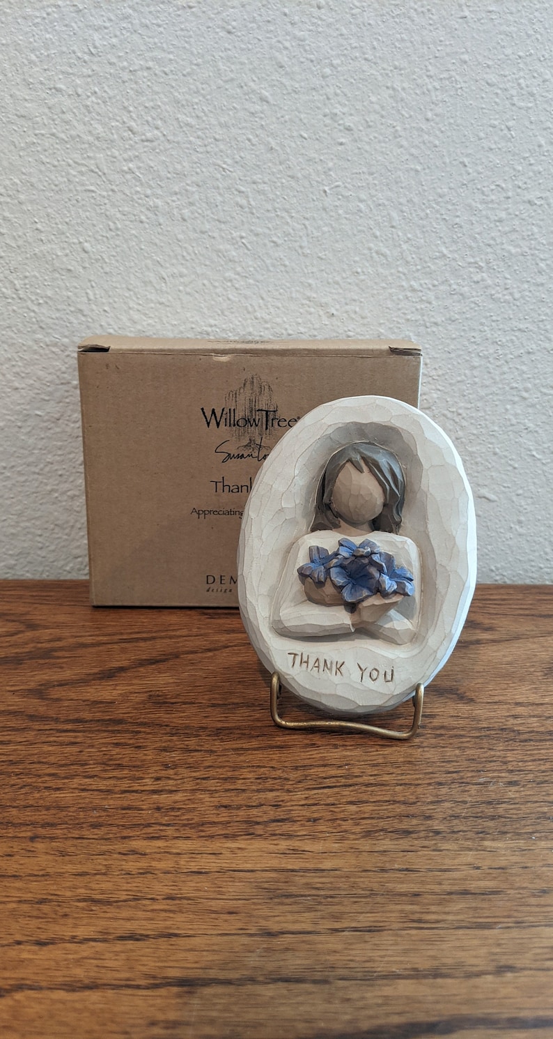 Puede incluir: Una figurita de madera tallada de una persona sosteniendo un ramo de flores azules. La figurita est&aacute; sobre un peque&ntilde;o soporte con las palabras "Gracias" inscritas en &eacute;l. La figurita est&aacute; hecha por Willow Tree by Susan Lordi.