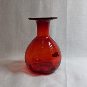 Pode incluir: Um pequeno vaso de vidro vermelho redondo com uma boca larga e um pescoço estreito.