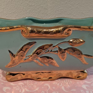 May include: A light blue ceramic planter with gold accents and a floral design. The planter has a scalloped rim and a wavy base.