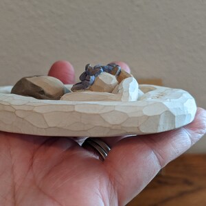 Puede incluir: Una peque&ntilde;a figura de madera blanca de una pareja durmiendo en una cama. La figura est&aacute; tallada con detalles y tiene un aspecto r&uacute;stico.