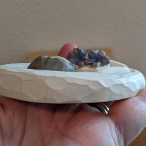 Puede incluir: Un peque&ntilde;o plato redondo de madera blanca con una superficie texturizada. El plato tiene tres peque&ntilde;os elementos decorativos tallados en la parte superior: una flor morada, una roca marr&oacute;n y una roca blanca.