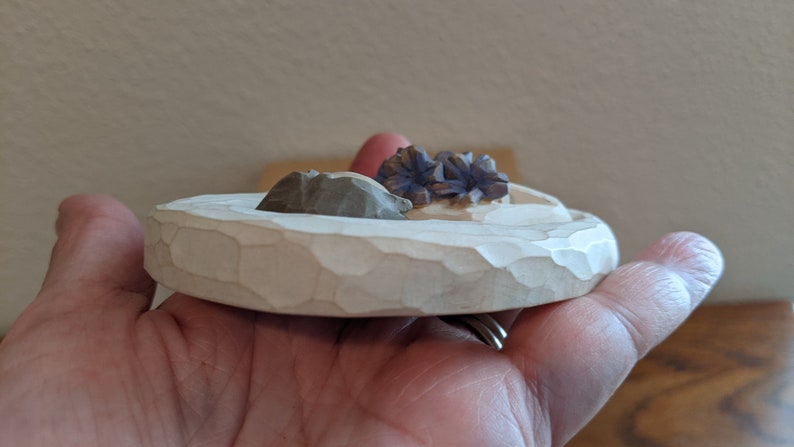 Puede incluir: Un peque&ntilde;o plato redondo de madera blanca con una superficie texturizada. El plato tiene tres peque&ntilde;os elementos decorativos tallados en la parte superior: una flor morada, una roca marr&oacute;n y una roca blanca.