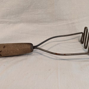 Puede incluir: Un machacador de patatas vintage de metal con mango de madera. El machacador tiene un diseño de alambre en espiral y el mango es marrón.