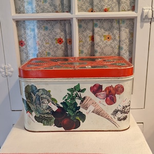 May include: Vintage red and white metal storage container with a floral design. The container has a hinged lid and is labeled with the names of different vegetables: 'Sweet Corn', 'Beets', 'Parsnip', 'Red Onion', and 'Garlic'.