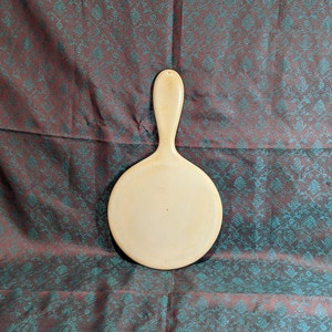 May include: Ivory-colored hand mirror with a long handle and a round mirror surface. The handle has a small hole at the top. The mirror is set against a patterned background with teal and burgundy colors.