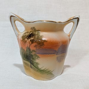 May include: A small, antique vase with two handles and a landscape painting. The vase features a cream-colored base with a painted scene of a tree, water, and a sunset in shades of orange, brown, and green. The handles and rim are trimmed with gold.