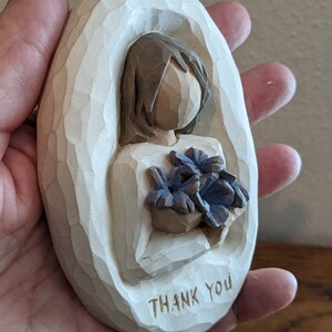 Puede incluir: Figurilla de madera blanca de forma ovalada que representa a una persona sosteniendo un ramo de flores azules. La figura tiene las palabras "Thank You" talladas en la parte inferior.