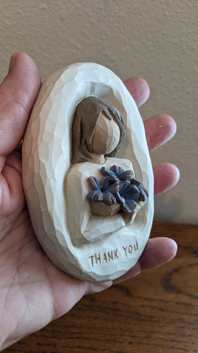Puede incluir: Figurilla de madera blanca de forma ovalada que representa a una persona sosteniendo un ramo de flores azules. La figura tiene las palabras "Thank You" talladas en la parte inferior.
