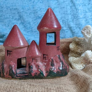 May include: A ceramic castle figurine with a pink and green glaze. The castle has four towers and is decorated with black lines. The figurine is sitting on a brown surface.