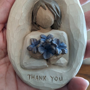 Puede incluir: Una placa ovalada de madera con una imagen tallada de una persona sosteniendo un ramo de flores azules. La placa tiene las palabras "Gracias" talladas en la parte inferior.