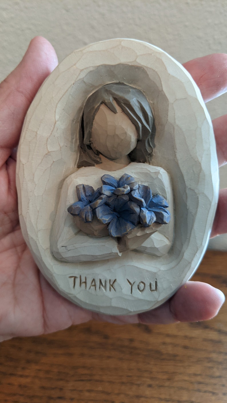 Puede incluir: Una placa ovalada de madera con una imagen tallada de una persona sosteniendo un ramo de flores azules. La placa tiene las palabras "Gracias" talladas en la parte inferior.