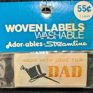 May include: A pack of cream-coloured woven labels with gold text reading "MADE WITH LOVE FOR DAD" and a black top hat illustration. The blue packaging displays "WOVEN LABELS WASHABLE" and "Ador-ables by Streamline".