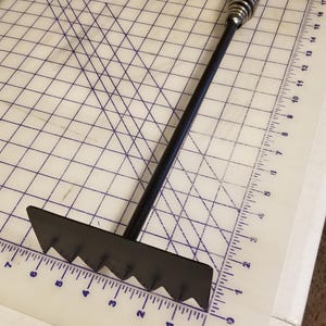 Peut inclure: Un râteau en métal noir avec un long manche et une poignée à ressort. Le râteau a une bordure dentelée et est conçu pour nettoyer un foyer.