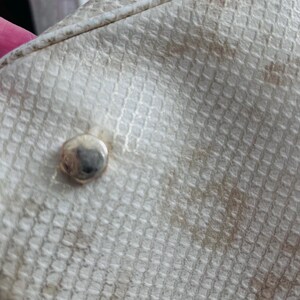 Pu&ograve; includere: Primo piano di una sezione di una borsa strutturata bianco sporco. La borsa ha un motivo quadrato ripetuto e un borchia color argento. Macchie marroni sono visibili sulla superficie della borsa. La borsa sembra essere fatta di un materiale sintetico.