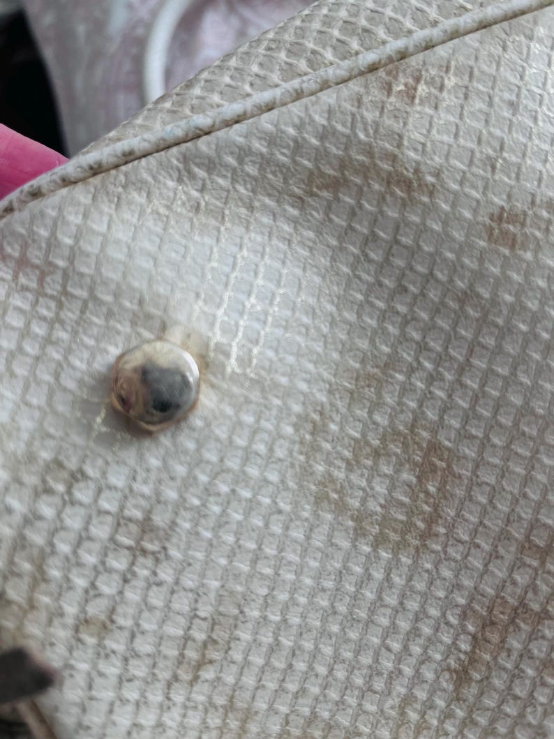 Pu&ograve; includere: Primo piano di una sezione di una borsa strutturata bianco sporco. La borsa ha un motivo quadrato ripetuto e un borchia color argento. Macchie marroni sono visibili sulla superficie della borsa. La borsa sembra essere fatta di un materiale sintetico.
