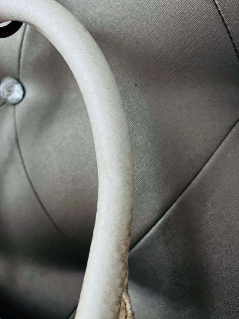 Pu&ograve; includere: Primo piano di un manico curvo bianco sporco con un motivo strutturato a scaglie. Il manico sembra far parte di una borsa, su uno sfondo grigio strutturato.