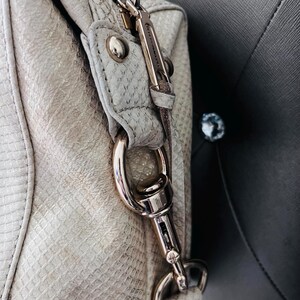 Pu&ograve; includere: Primo piano di una borsa grigio chiaro con un motivo strutturato simile alla pelle di serpente. La borsa presenta elementi metallici color argento, tra cui una chiusura, una cerniera e rivetti decorativi. La tracolla della borsa &egrave; fissata con un moschettone in metallo argentato.