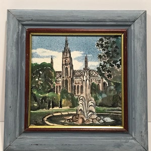 May include: Framed watercolor painting of a cathedral with a fountain in the foreground. The painting features a tall, ornate building with multiple spires, surrounded by trees and a circular fountain. The frame is a distressed blue with a brown inner border.