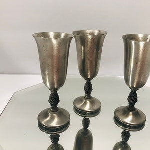 May include: Three silver goblets with a decorative design on the stem. The goblets are on a mirrored surface.