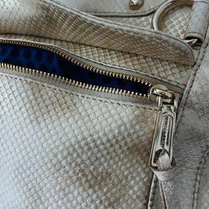 Pu&ograve; includere: Primo piano di una borsa argentata metallizzata con una texture a pelle di serpente. La borsa presenta una cerniera dorata con il nome del marchio "Rebecca Minkoff" sul tirazip. La fodera interna &egrave; in tessuto blu con un motivo a pois.