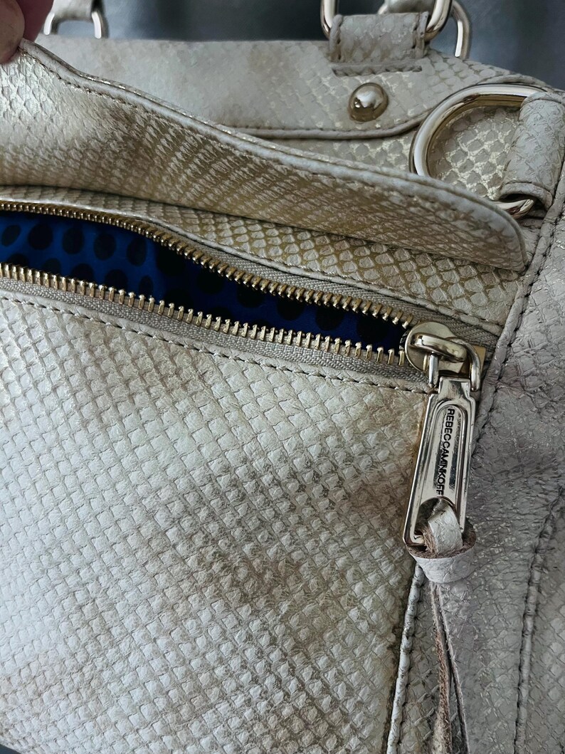Pu&ograve; includere: Primo piano di una borsa argentata metallizzata con una texture a pelle di serpente. La borsa presenta una cerniera dorata con il nome del marchio "Rebecca Minkoff" sul tirazip. La fodera interna &egrave; in tessuto blu con un motivo a pois.