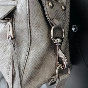 Pu&ograve; includere: Primo piano di una borsa grigio chiaro con motivo a pelle di serpente. La borsa presenta hardware color argento, tra cui cerniere, chiusure e anelli. Il marchio "REBECCAMINKOFF" &egrave; visibile su un tirazip.