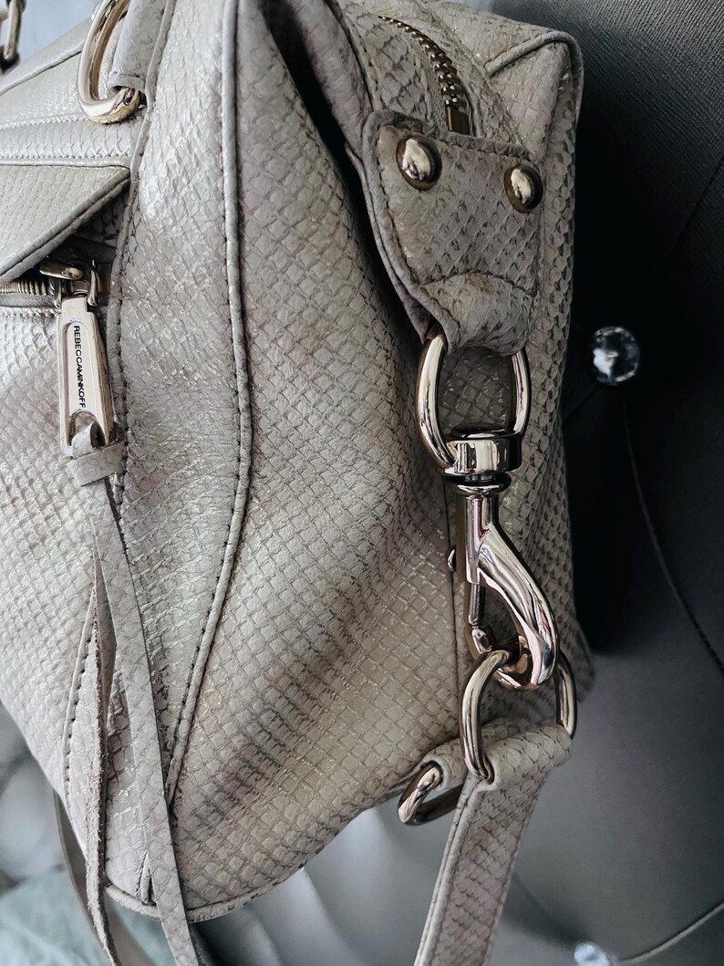 Pu&ograve; includere: Primo piano di una borsa grigio chiaro con motivo a pelle di serpente. La borsa presenta hardware color argento, tra cui cerniere, chiusure e anelli. Il marchio "REBECCAMINKOFF" &egrave; visibile su un tirazip.