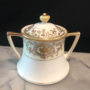 May include: A white porcelain sugar bowl with a lid, featuring gold and black floral designs. The bowl has two handles and a gold rim.