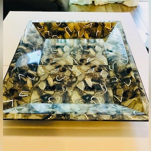 May include: A rectangular decorative tray with a mosaic pattern of brown, gray, and white stones. The tray has a clear, beveled edge and a glossy finish. The tray is designed for decorative purposes.