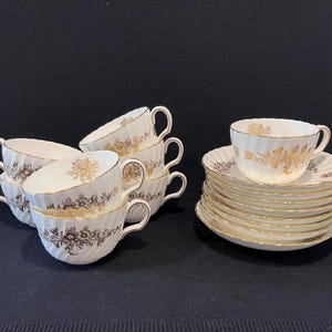 May include: A collection of white teacups and saucers with gold floral designs. The teacups have a curved shape and are stacked together. The saucers are stacked separately. The rims of the cups and saucers are trimmed with gold.