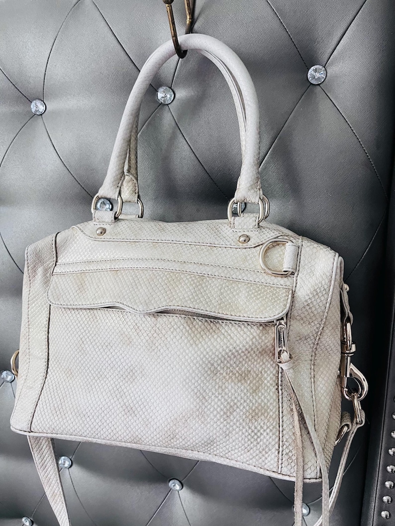 Pu&ograve; includere: Una borsa grigio chiaro con un motivo a pelle di serpente, con un manico superiore e una tracolla staccabile. La borsa ha una tasca frontale con cerniera e hardware color argento. La borsa &egrave; appesa a un gancio.