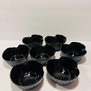 May include: A set of six glossy black bowls with a textured, basket-weave pattern on the exterior. Each bowl features a scalloped, wavy rim and a smooth, reflective interior surface. The bowls are arranged in a cluster on a plain white surface.