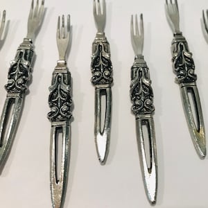 May include: Set of eight silver-toned, ornate, decorative forks with a detailed floral design. The forks are approximately 4 inches long.