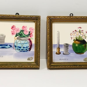 May include: Two framed watercolor paintings by M. Angelo. The first painting depicts a blue and white vase with pink flowers and a blue plate with a glass. The second painting shows a green vase with white flowers, a candle, and a silver cup. Both paintings have a light blue background.