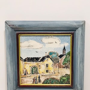 May include: A framed painting of a small village scene with a yellow building, a church steeple, and figures walking on a path. The painting is done in a watercolor style with a textured background.